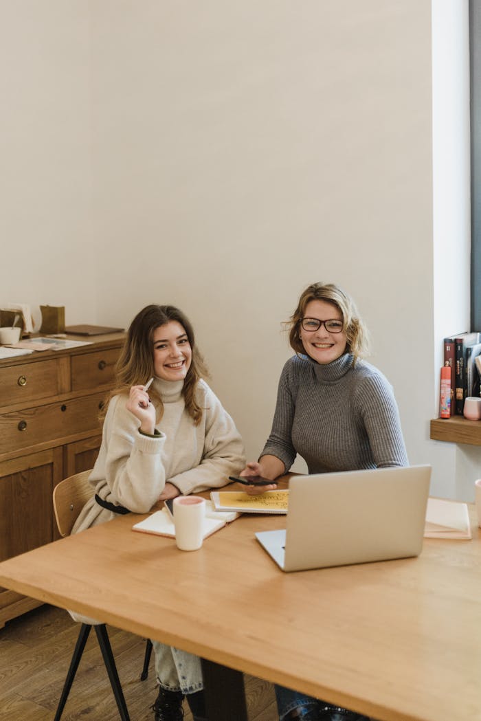 Two women smiling and working together at a desk in a modern office environment.